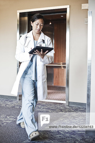 Asian woman doctor walking in a hospital hallway while working on a notebook computer.