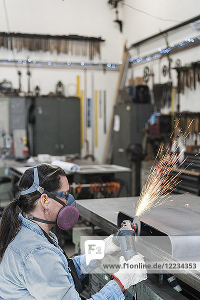 Frau mit Schutzbrille und Staubmaske steht in Metallwerkstatt  benutzt Schleifmaschine  Funkenflug.