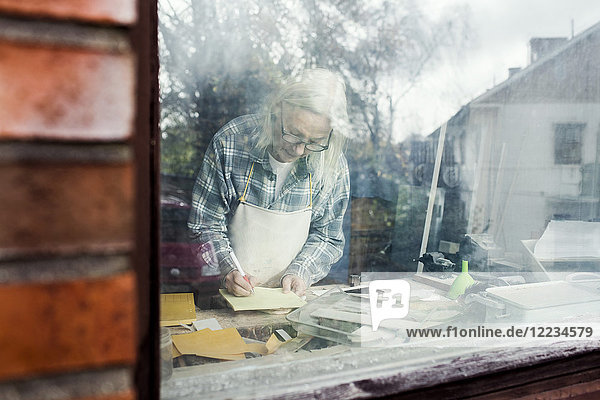 Senior Handwerker beim Schreiben auf Papier durch Glasfenster gesehen