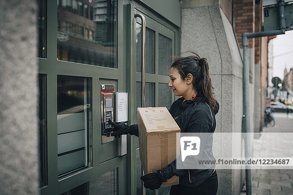 Female messenger ringing intercom on closed door while carrying box on sidewalk