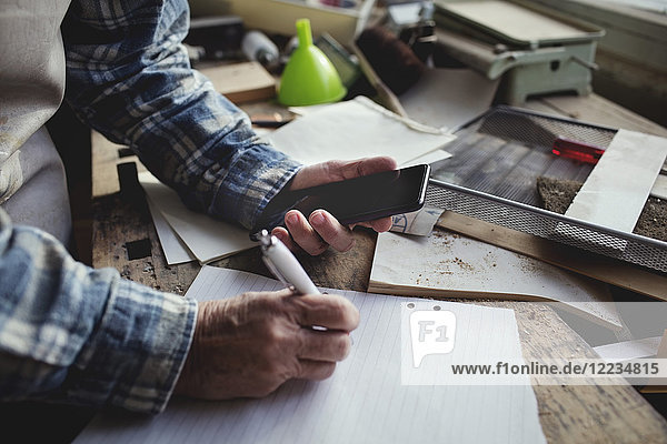 Cropped image of owner holding mobile phone while writing on paper at workbench