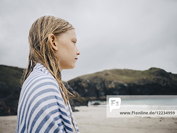 Seitenansicht des blonden Mädchens  eingewickelt in ein gestreiftes blaues Handtuch am Strand gegen den Himmel.