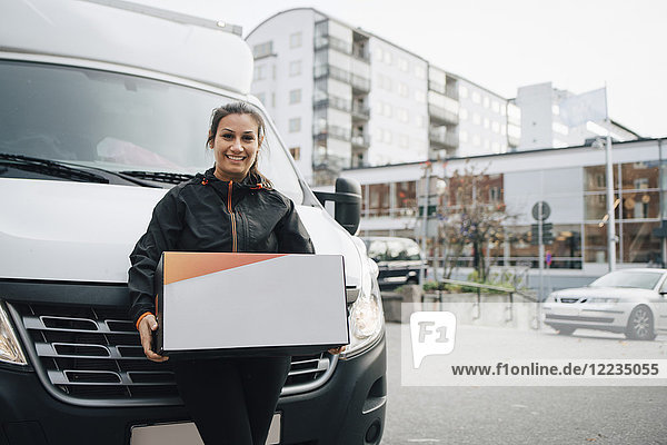 Porträt einer lächelnden Arbeiterin  die eine Kiste trägt  während sie gegen einen Lieferwagen in der Stadt steht.