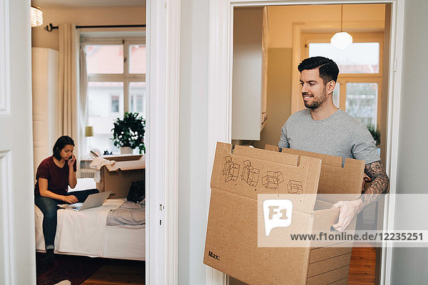 Lächelnder Mann trägt Box von der Tür zur Frau mit Laptop im Schlafzimmer
