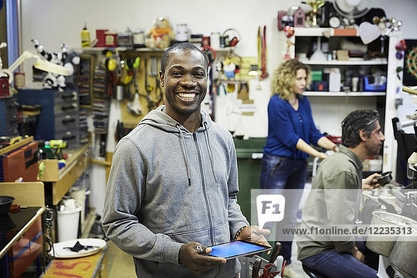 Porträt eines lächelnden Freiwilligen mit digitalem Tablett und Kollegen  die in der Werkstatt arbeiten.