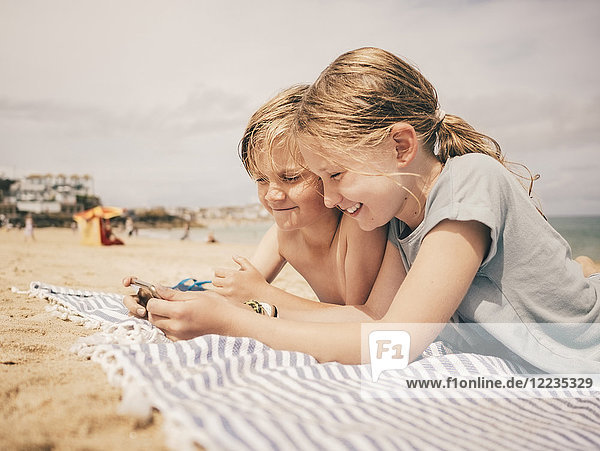 Happy siblings sharing smart phone while lying on towel at beach against sky