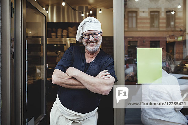 Smiling male baker standing with arms crossed at entrance of bakery