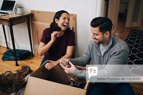 Lachendes Paar beim Auspacken der Pappschachtel im Sitzen auf dem Boden zu Hause