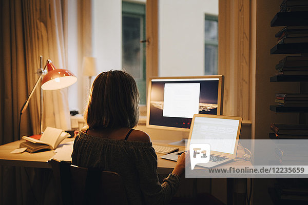 Rückansicht des Mädchens mit Laptop und Computer im Sitzen am beleuchteten Schreibtisch