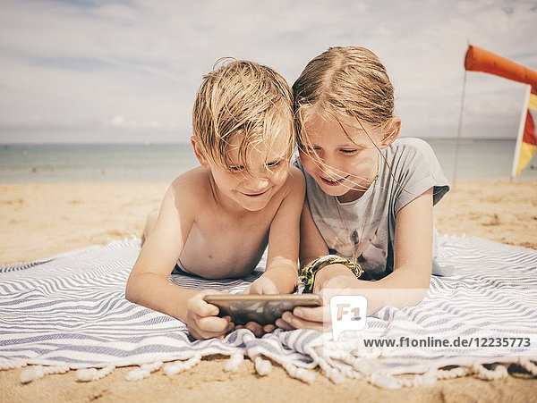 Lächelnde Geschwister teilen sich ein Smartphone  während sie auf einem Handtuch am Strand gegen den Himmel liegen.