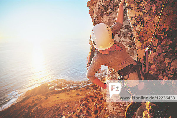 Female rock climber reaching for clip above sunny ocean
