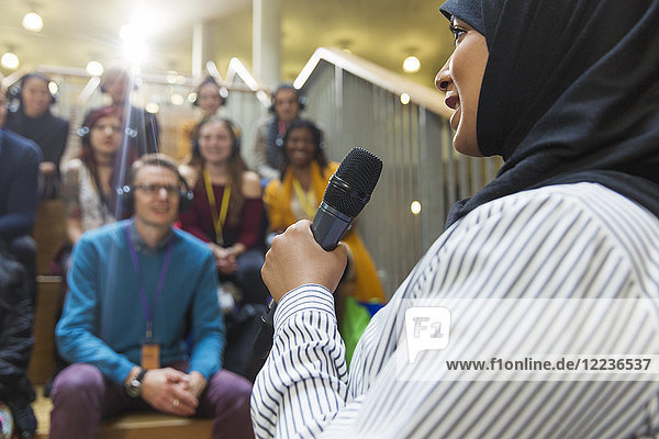 Lächelnde Geschäftsfrau im Hijab  die mit Mikrofon vor Publikum spricht.