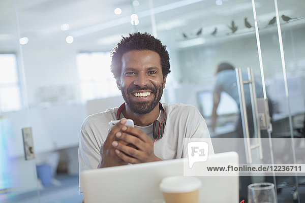 Porträt eines lächelnden  selbstbewussten  kreativen Geschäftsmannes  der ein Smartphone am Laptop im Büro benutzt