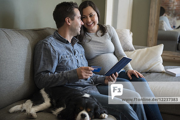 Mid adult couple shopping online in living room  sitting on sofa with puppy