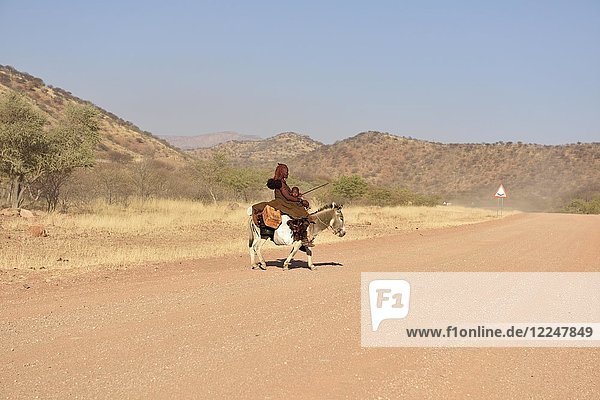 Junge  verheiratete Himbafrau reitet mit einem kleinen Kind auf einem Esel  Kaokoveld  Namibia  Afrika