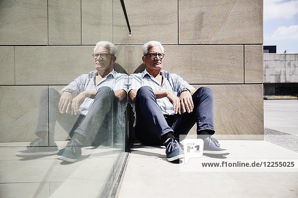 Grey-haired senior on the floor in front of a wall and reflected in a glass pane  Cologne  North Rhine-Westphalia  Germany  Europe