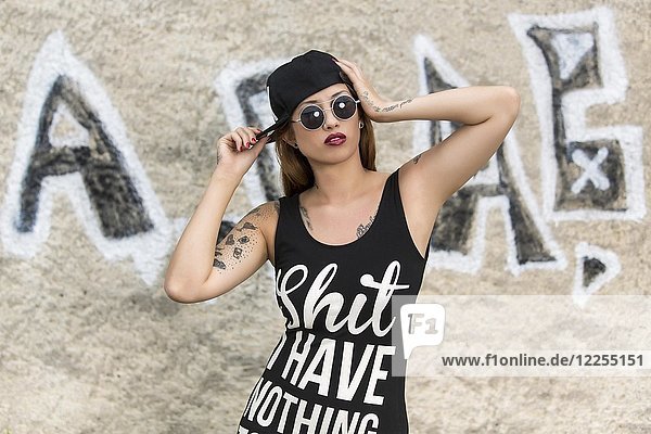 Young tattooed woman in a shirt and with cap in front of the graffiti wall