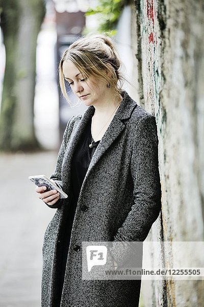 Junge Frau lehnt an einer Wand und schaut auf ihr Smartphone  Köln  Nordrhein-Westfalen  Deutschland  Europa