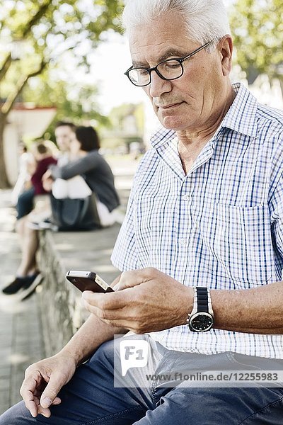 Grey-haired senior sits on a wall with his smartphone in his hand  Cologne  North Rhine-Westphalia  Germany  Europe