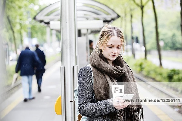 Junge Frau wartet mit ihrem Smartphone in der Hand an einer S-Bahn-Station  Deutschland  Europa