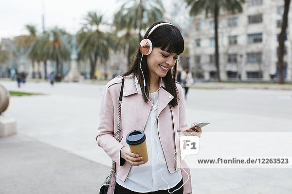 Spanien  Barcelona  lächelnde Frau mit Kaffee  Handy und Kopfhörer in der Stadt