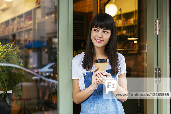 Lächelnde Frau steht vor der Eingangstür eines Ladens mit Kaffee zum Mitnehmen.