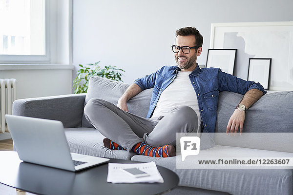 Relaxed smiling man sitting on sofa looking at laptop