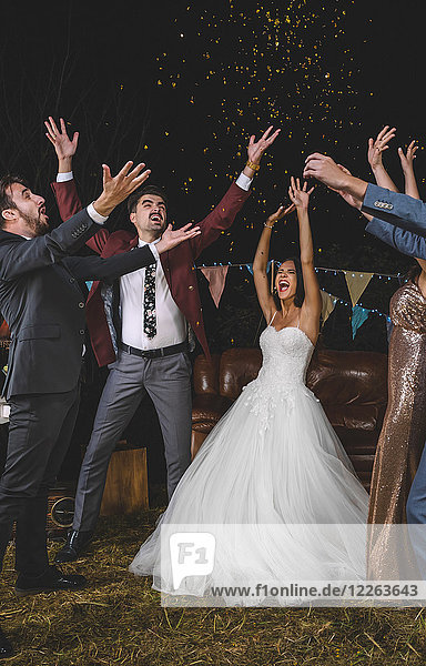 Fröhliches Hochzeitspaar und Freunde  die ihre Arme heben  während Konfetti auf einer Nachtparty im Freien über den Kopf fallen.