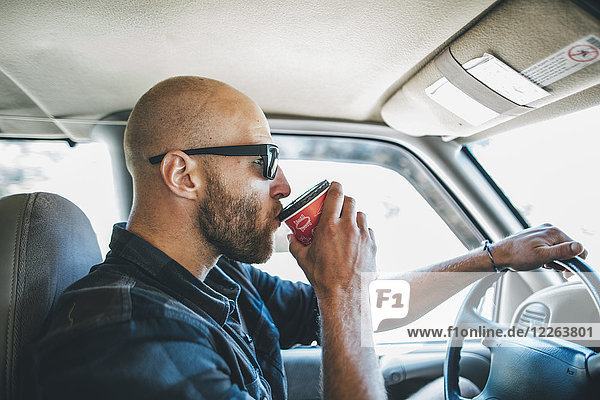Junger Mann mit Sonnenbrille und Bart auf einem Roadtrip mit Takeaway-Getränk