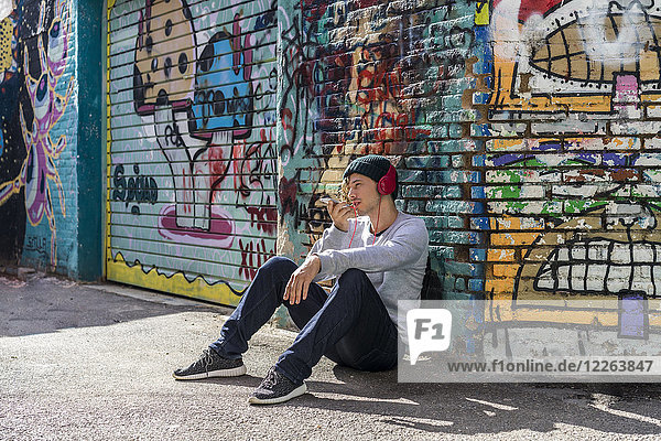 Young man with headphones sitting in front of graffiti wall using cell phone