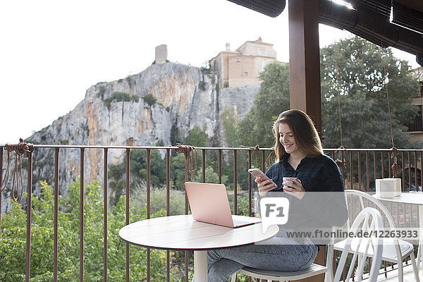 Spanien  Alquezar  lächelnde junge Frau auf der Terrasse sitzend mit Laptop per Handy