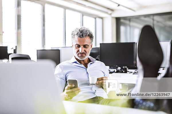 Relaxed mature businessman sitting at desk in office using cell phone and drinking coffee