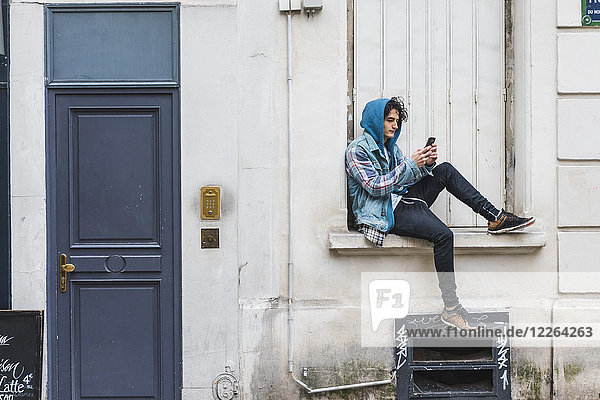 Junger Mann mit Smartphone auf der Fensterbank sitzend