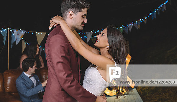 Romantic wedding couple embracing on a night field party with their friends in the background
