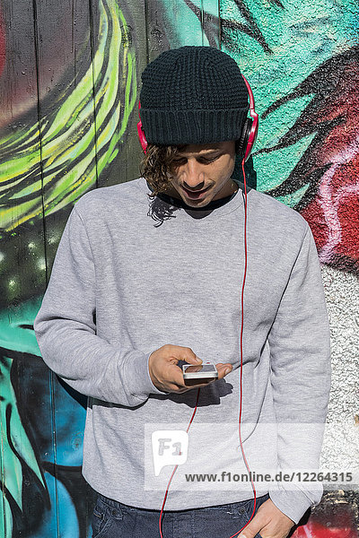 Junger Mann vor der Graffiti-Wand hört Musik mit Kopfhörern  während er ein Handy benutzt.