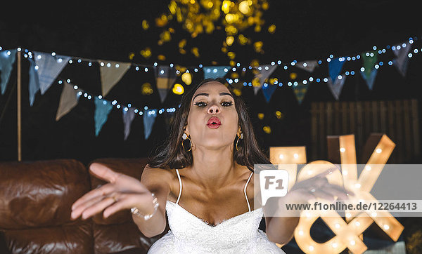 Porträt einer glücklichen Braut  die Konfetti auf einer Nachtparty im Freien bläst.
