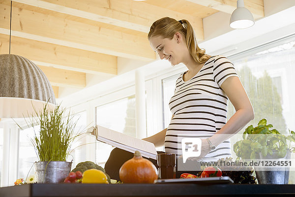 Lächelnde schwangere Frau liest Buch in der Küche zu Hause