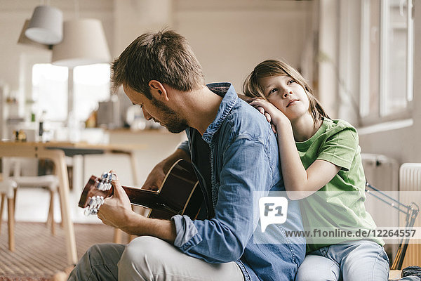 Sohn lehnt sich an Vater  der zu Hause Gitarre spielt.