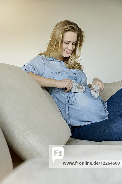 Lächelnde schwangere Frau sitzt auf der Couch und hält Babyschuhe.