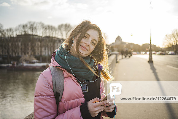 Paris  Frankreich  Portrait einer lächelnden jungen Frau beim Musikhören mit Kopfhörern und Smartphone