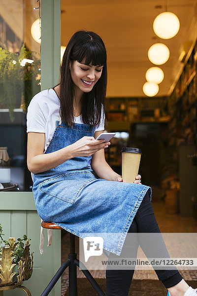 Smiling woman sitting on stool using cell phone at entrance door of a store
