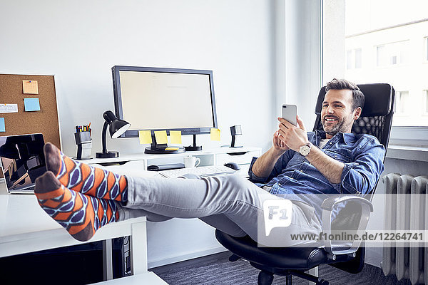 Entspannter Mann sitzt am Schreibtisch im Büro mit dem Handy