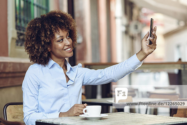Lächelnde Frau mit Afro-Frisur sitzend im Outdoor-Café mit einem Selfie