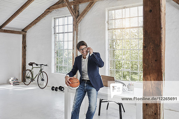 Lächelnder Geschäftsmann mit Basketball am Handy im Penthouse