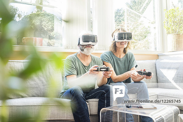 Ein reifes Paar das zu Hause auf der Couch sitzt und eine VR-Brille trägt und ein Videospiel spielt.