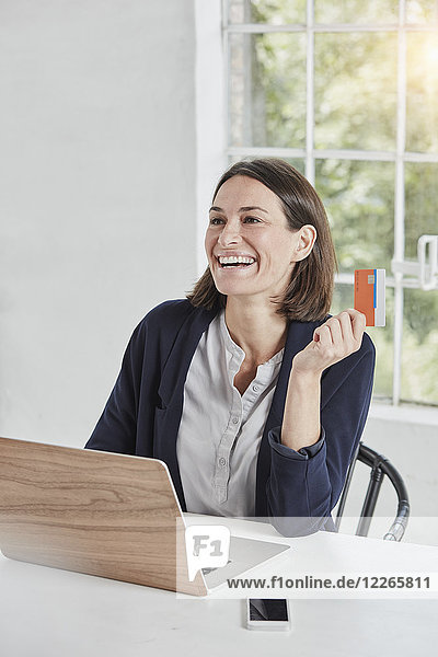Laughing businesswoman using laptop on desk holding card