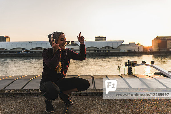 Fröhliche sportliche junge Frau beim Musikhören am Flussufer in der Stadt bei Sonnenuntergang
