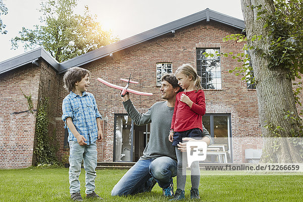 Vater mit zwei Kindern  die mit einem Spielzeugflugzeug im Garten ihres Hauses spielen.
