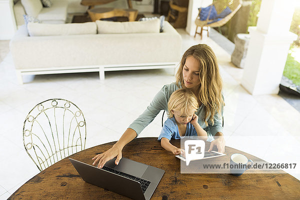 Boy sitting on his mother's lap and looking at a tablet while his mother is working on a laptop