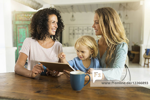 Zwei lächelnde Frauen mit einem Kind mit Tablette am Küchentisch